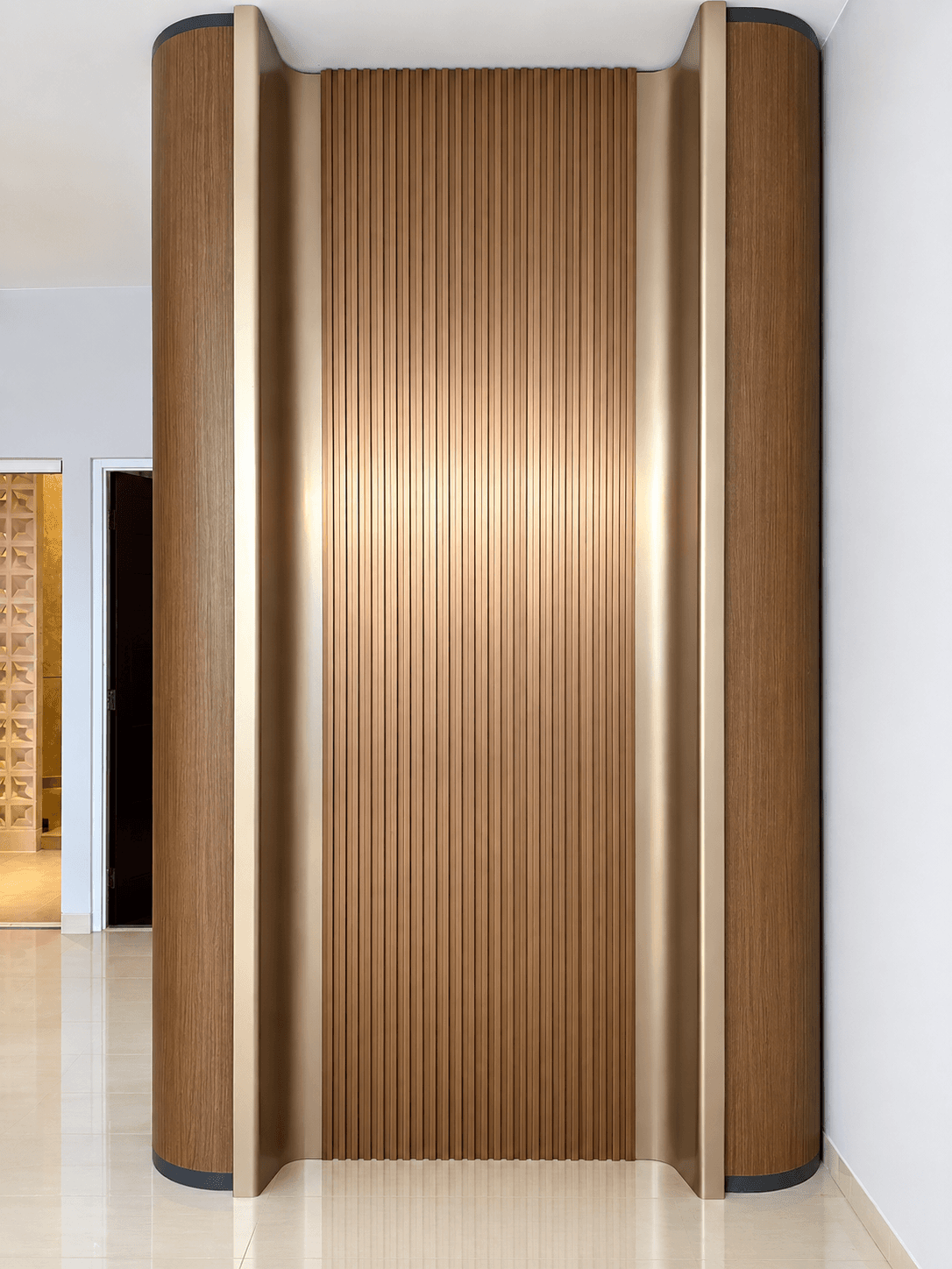Dinding foyer dengan panel kayu vertikal di tengah, diapit wallpanel kayu melengkung berbingkai rosegold dan lampu tersembunyi
