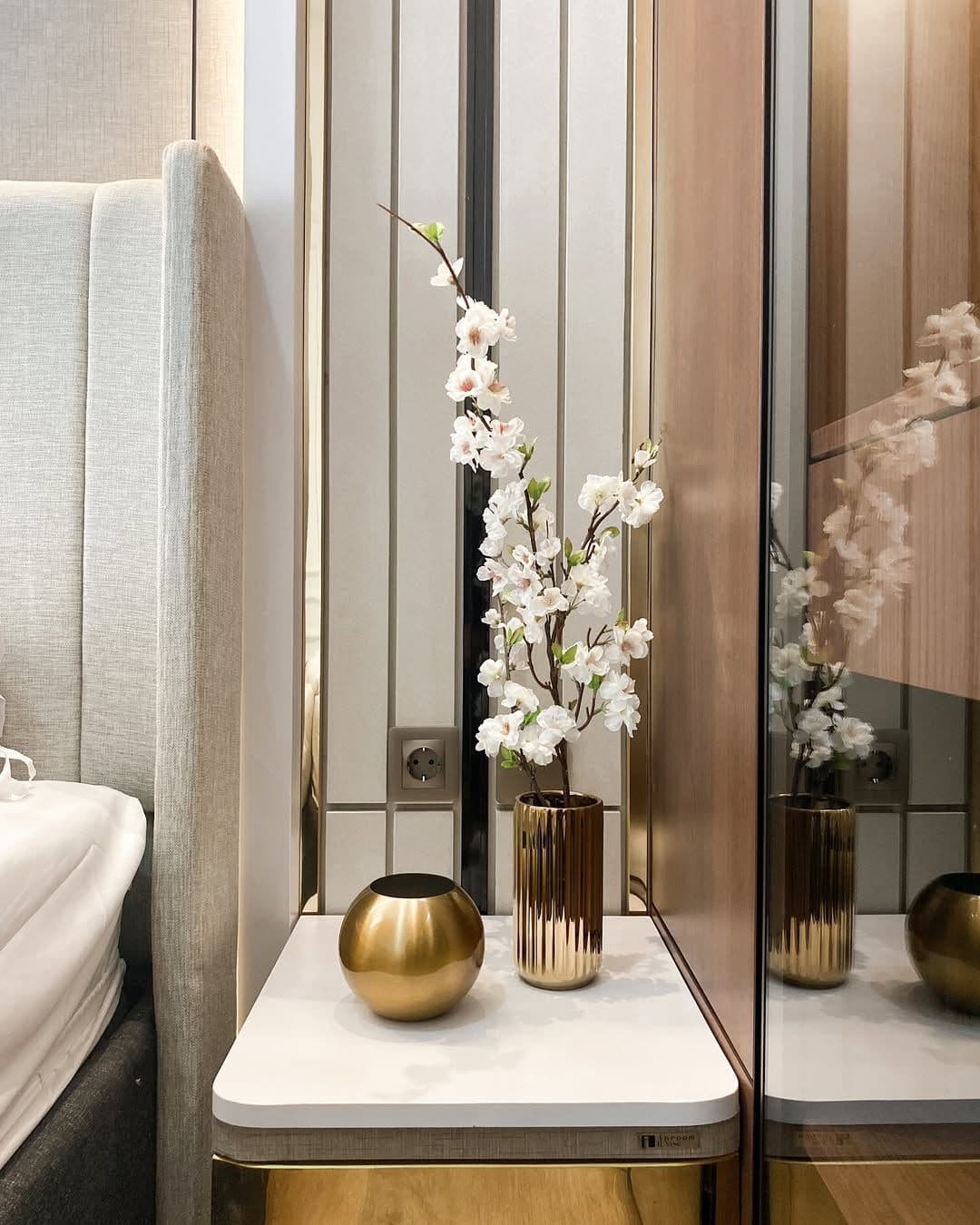 White marble bedside table with a brass vase, against a cream panelled wall lined with gold stainless and a mirror